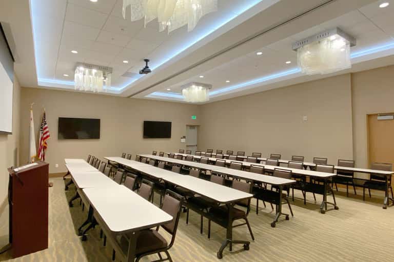 Spacious ballroom with long conference tables, chairs, ceiling fixtures, and flat-screen displays