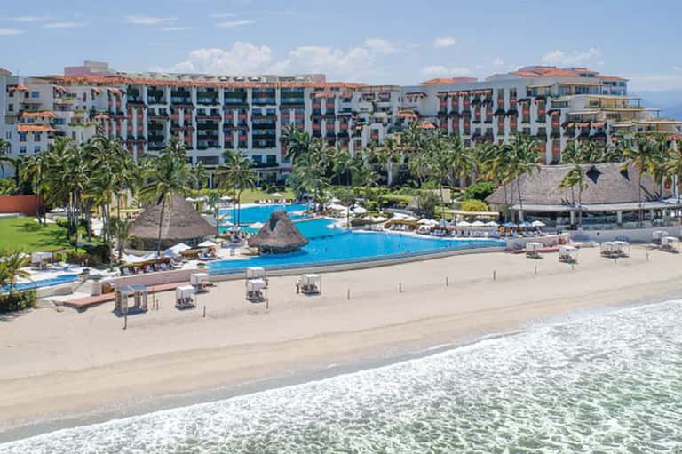 Large beachfront resort with tall residential buildings, multiple pools, thatched cabanas, and sandy beach