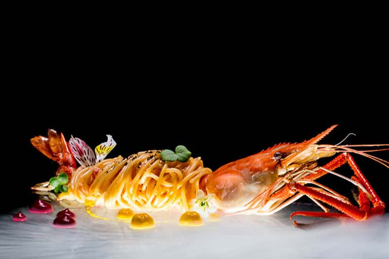 Elegant plated lobster pasta dish with decorative chocolate elements and colorful garnishes on white plate