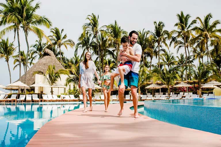 Family of four jogging on a dock between two pools at a tropical resort with palm trees and thatched roofs
