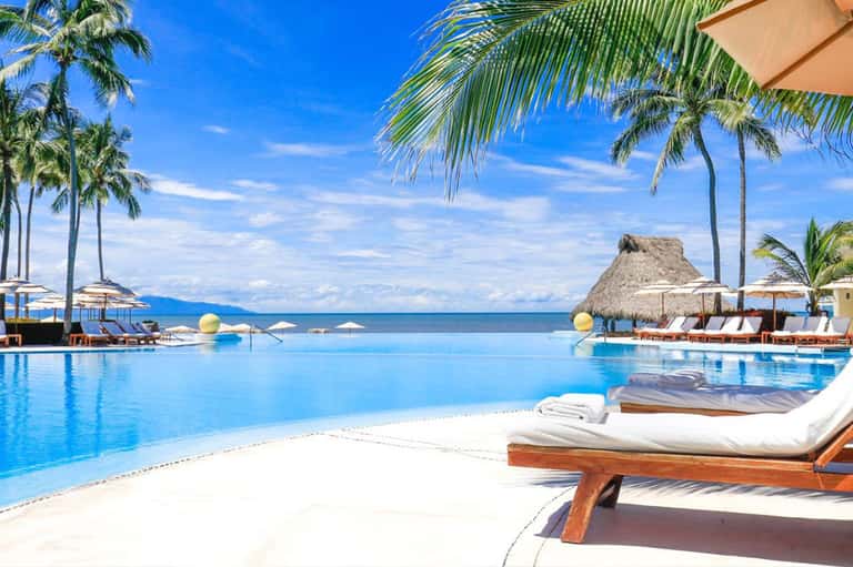 Luxury infinity pool blending with ocean horizon, white lounge beds, thatched umbrella, and palm trees at sunset