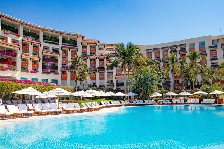 Large resort pool with turquoise water, white lounge chairs, colorful flower-draped building facade