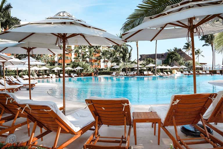 Resort pool deck with turquoise water, lounge chairs, white umbrellas, palm trees, and colorful colonial-style buildings