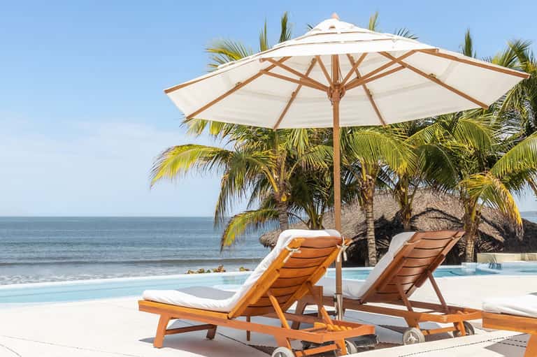 Beachfront lounge chairs with wooden frames and cream cushions under large umbrella, palm trees, and ocean view