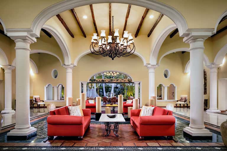 Grand hotel lobby with arched columns, crystal chandelier, red armchairs, and tropical garden views through glass doors