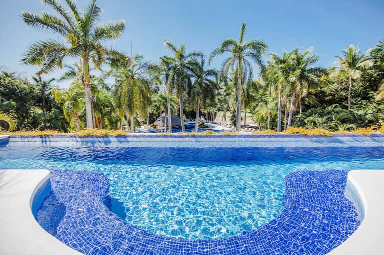 Resort swimming pool with blue mosaic tile and palm trees, clear sunny day with loungers visible