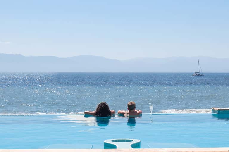 Infinity pool overlooking ocean with two swimmers, sailboat visible on water