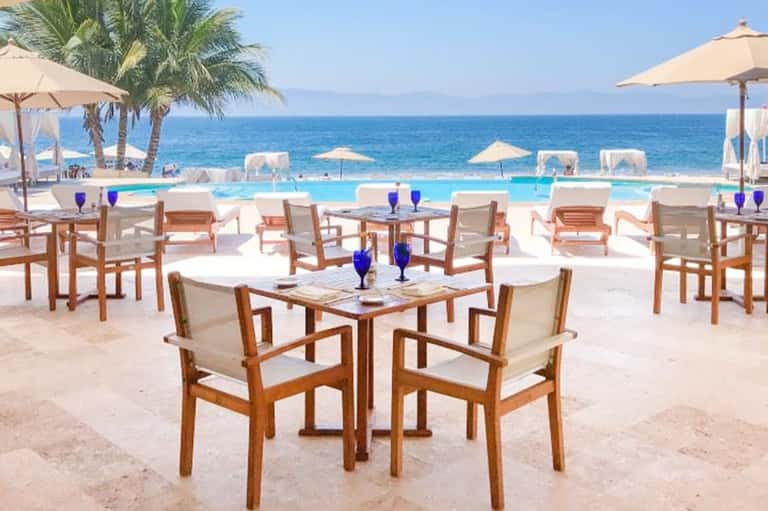 Beachfront resort dining area with wooden chairs, blue cocktails, pool, ocean view, and palm trees