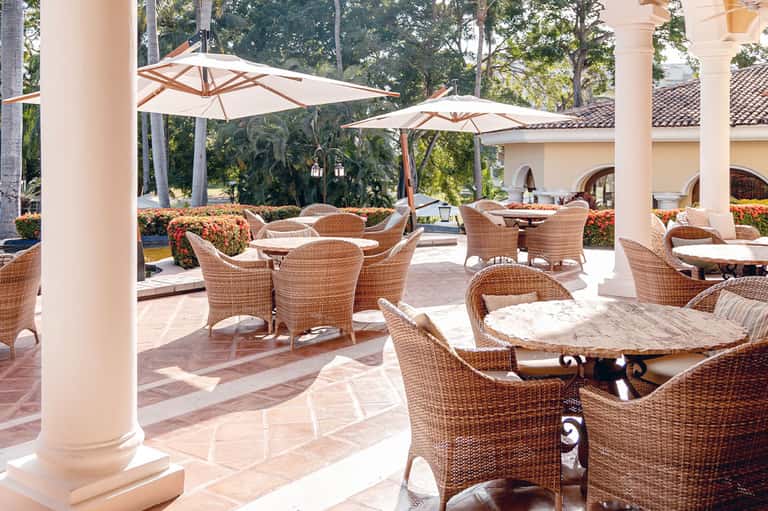 Elegant resort patio with wicker furniture, white umbrellas, columns, and lush landscaping