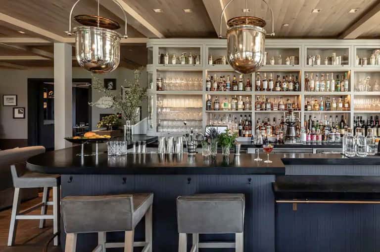 Modern bar with dark counter, pendant lighting, fully stocked shelves, and upholstered bar stools