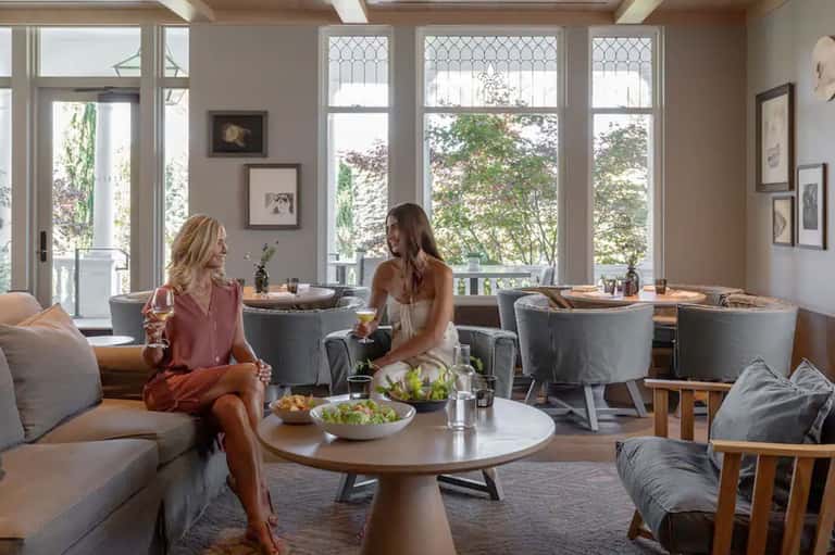 Two women enjoying drinks in an elegant lounge with floor-to-ceiling windows overlooking trees