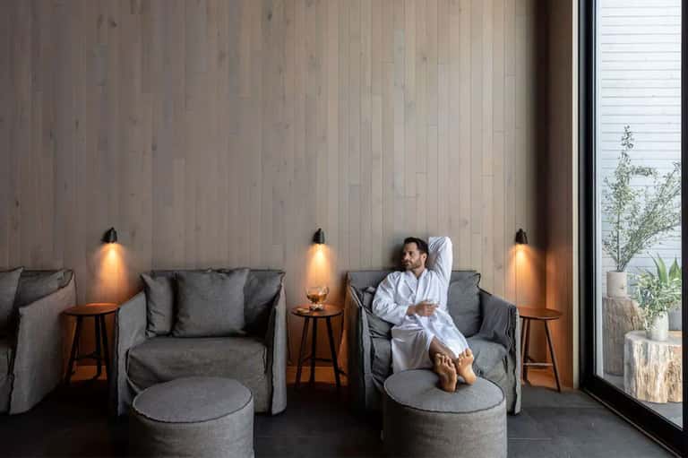 Spa relaxation area with gray armchairs, wooden side tables, pendant lights, and water view