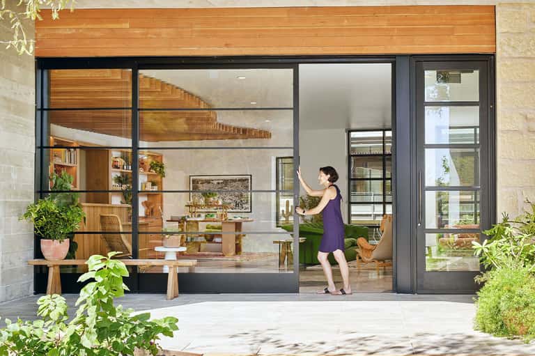 Woman opening large black-framed glass doors to a modern wooden interior with plants and shelving