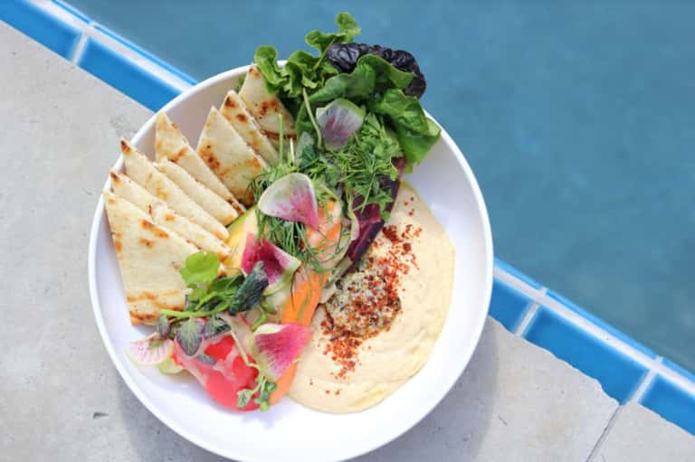 Elegant plated dish with hummus, fresh greens, colorful vegetables, pita bread, and spiced garnish by poolside
