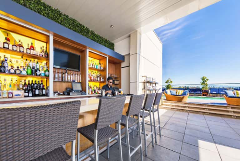 Rooftop bar with yellow-lit shelving, wicker bar seating, and waterfront views on a sunny day