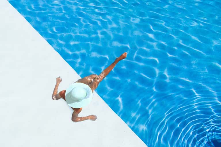 Person swimming in crystal-clear blue pool wearing white hat, overhead view