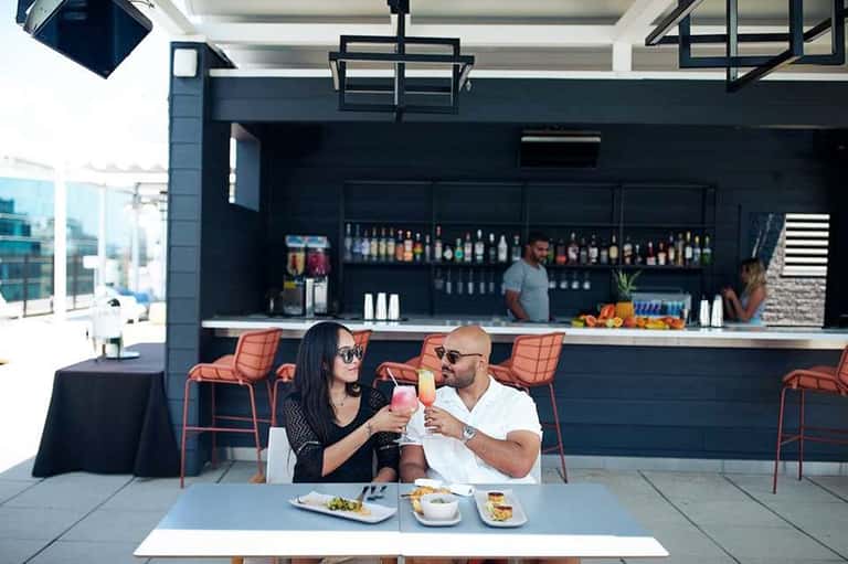 Contemporary rooftop bar with couple dining, dark counter, orange seating, and city skyline backdrop