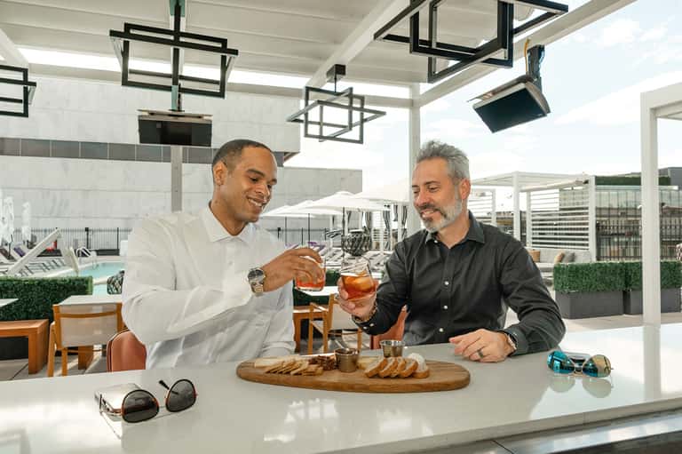 Two men enjoying drinks on modern rooftop bar overlooking pool with contemporary architecture
