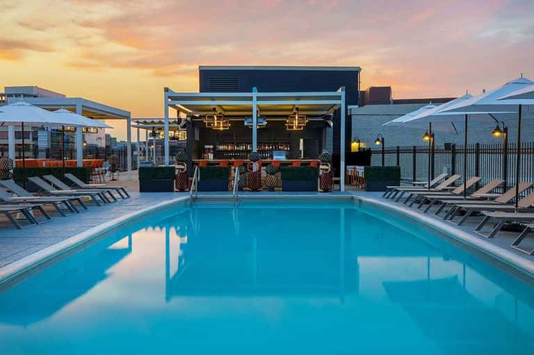 Resort pool at dusk with modern bar pavilion, lounge chairs, umbrellas, and sunset sky reflecting in water
