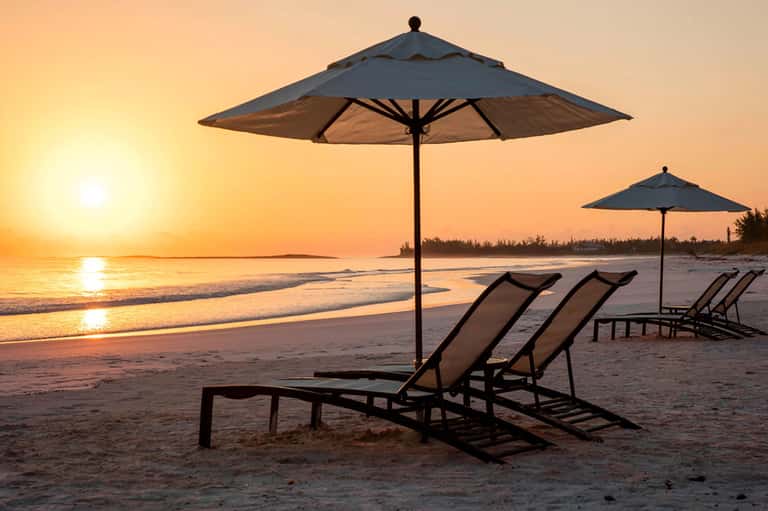 Beach sunset view with two lounge chairs under large umbrella on sandy shore at dusk