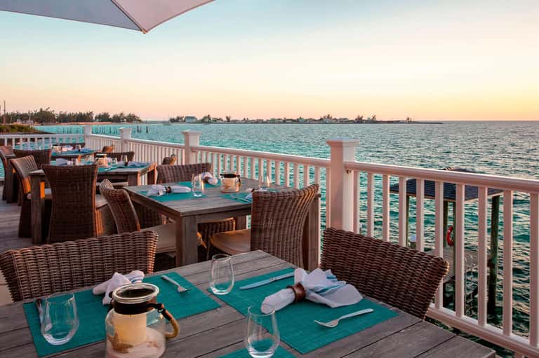 Waterfront dining deck with rattan chairs, turquoise placemats, and ocean sunset view
