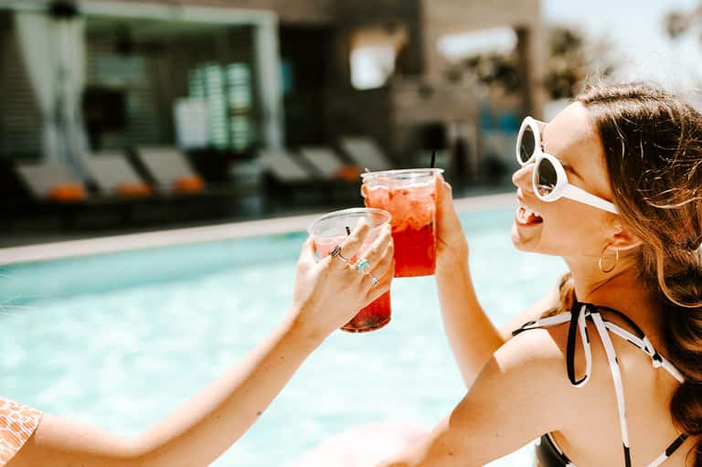 Woman in white sunglasses holding colorful drinks by pool lounge area