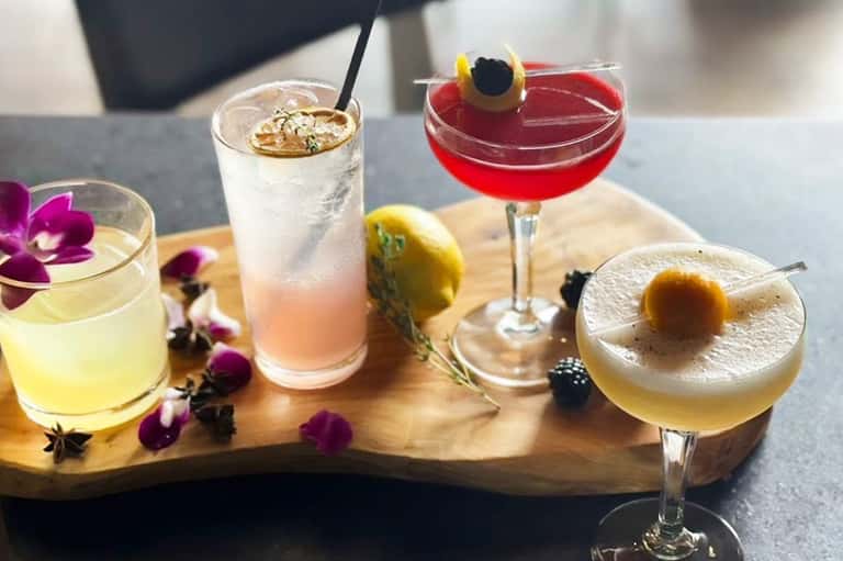 Four colorful cocktails on wooden board with purple flowers and citrus garnishes