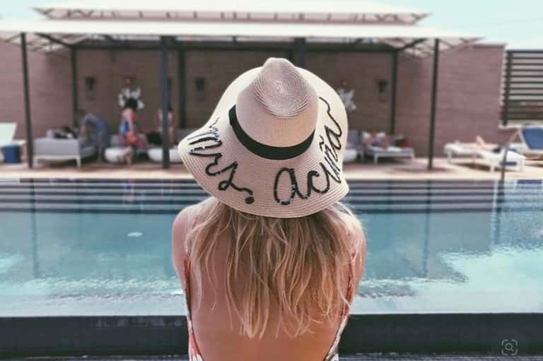 Woman in white sun hat sitting at edge of resort pool facing covered seating area
