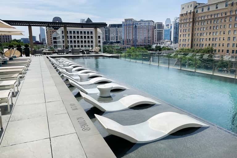 Modern white lounge chairs arranged along turquoise infinity pool with urban skyline and buildings