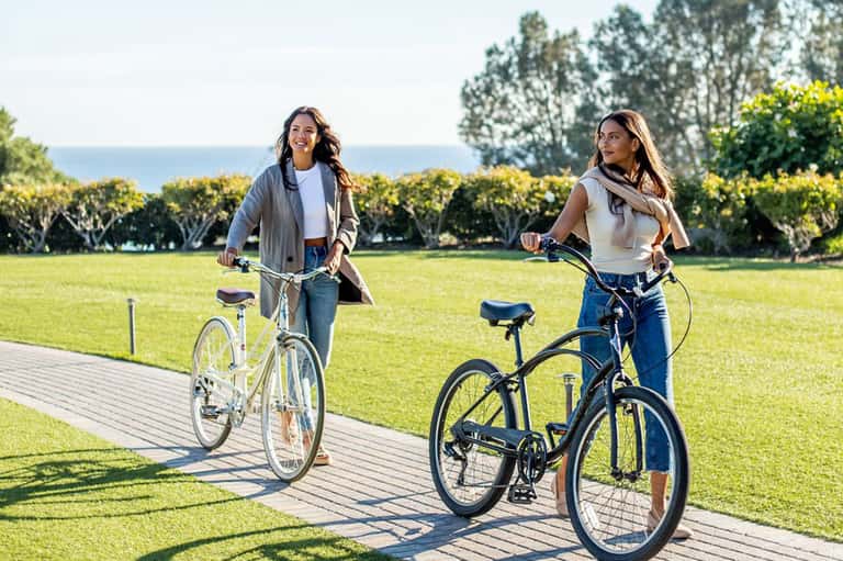 Two women riding bicycles on wooden boardwalk with ocean and trees visible in background