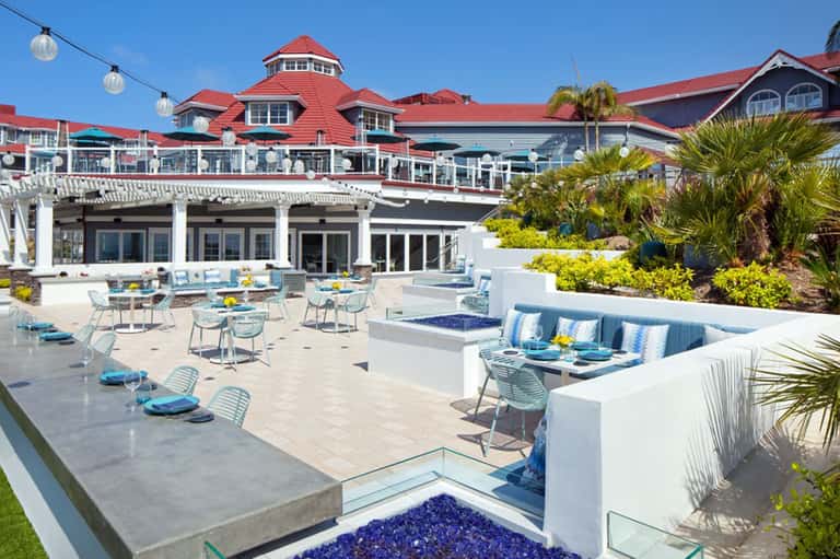Beachfront resort terrace with blue accents, white lounge seating, and red-roofed buildings