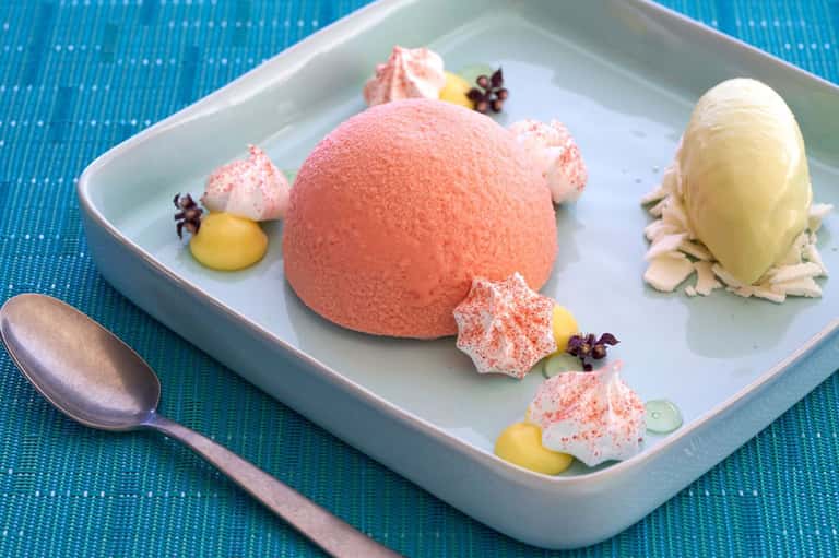 Gourmet dessert plate with pink mousse sphere, meringues, and yellow sauce on blue textured surface