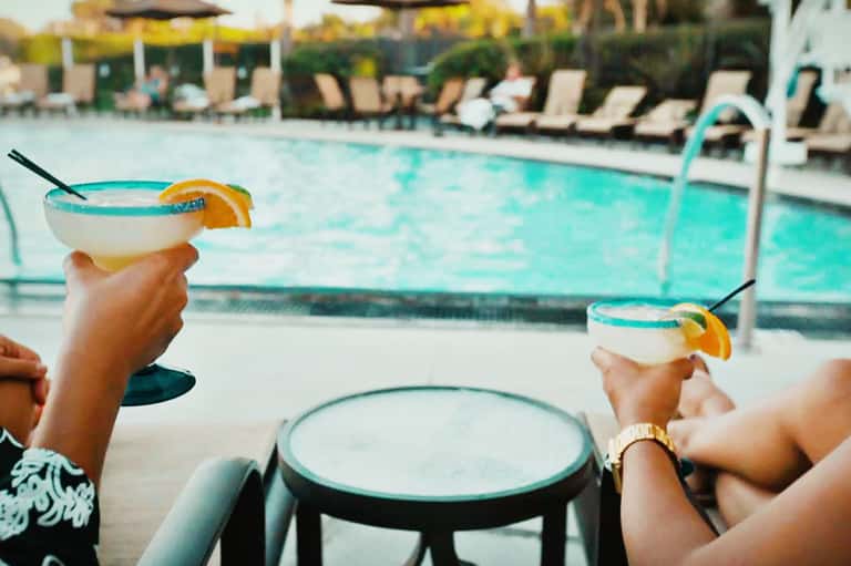 Two hands holding tropical cocktails with orange slices by resort pool with lounge chairs