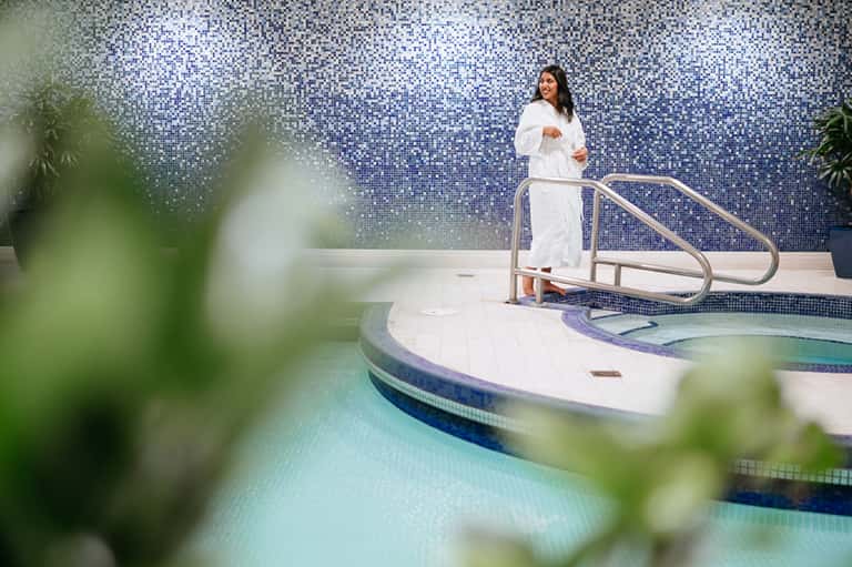 Woman in white robe standing by indoor spa pool with blue mosaic tile wall