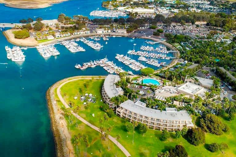 Aerial view of marina with white boats docked, curved waterfront building, and lush green grounds surrounded by blue water
