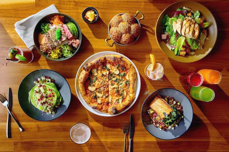 Overhead view of restaurant table with colorful dishes, pizza, salads, and fresh juices on wooden surface