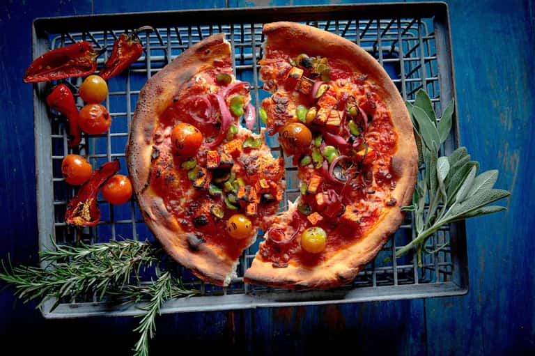 Wood-fired pizza with toppings and fresh herbs on a blue background with colorful presentation