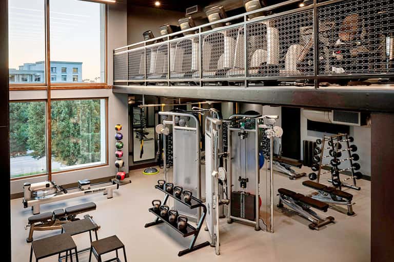 Modern hotel gym with weights, machines, large windows, and mezzanine level storage