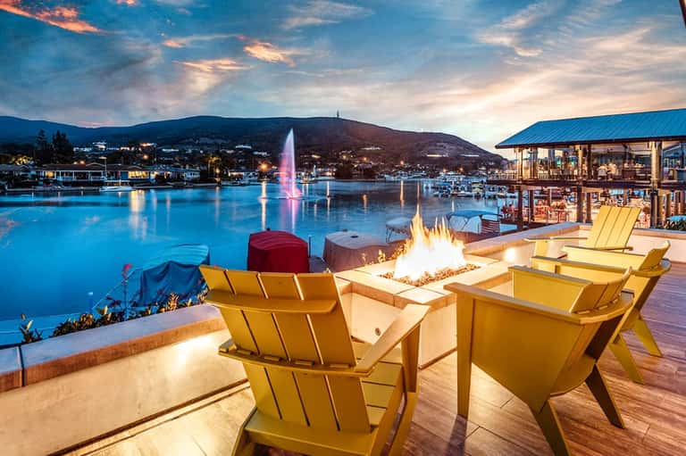 Yellow Adirondack chairs by fire pit overlooking illuminated fountain and mountains across blue lake at dusk