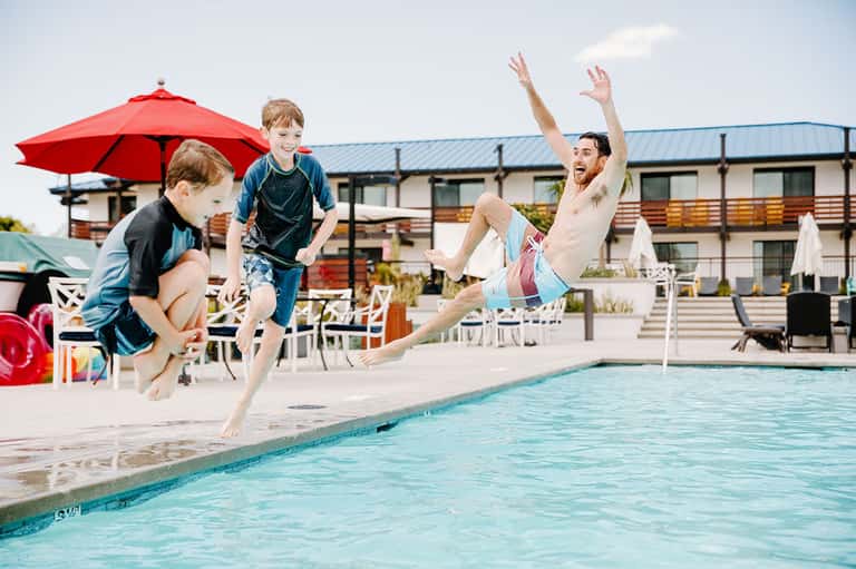 Family jumping into resort pool with red umbrella, modern building, and deck seating