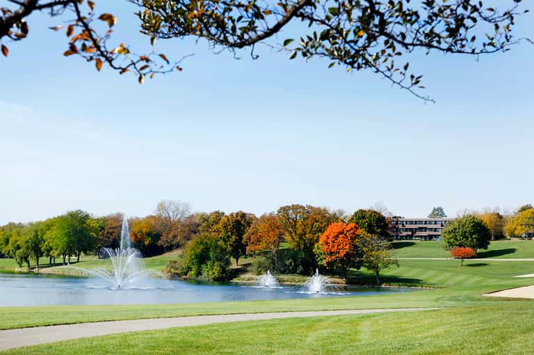 Golf course with manicured lawn, pond with fountains, fall foliage trees, and resort building in distance