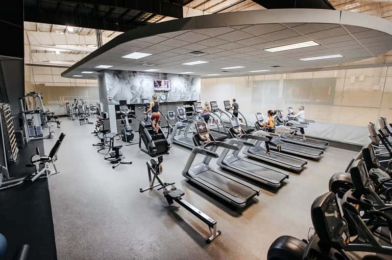 Large fitness studio with rows of treadmills, stationary bikes, and exercise equipment under industrial ceiling