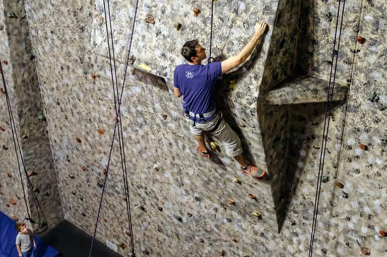 Man rock climbing indoor wall wearing blue shirt with rope assistance and holds scattered across grey textured surface