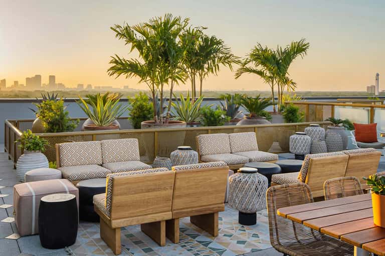 Sunset rooftop lounge with geometric patterned seating, palm trees, and waterfront skyline view