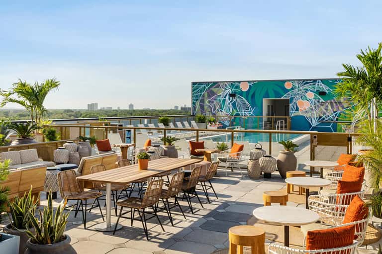 Rooftop terrace with colorful seating, wooden tables, city skyline views, and mural accent wall