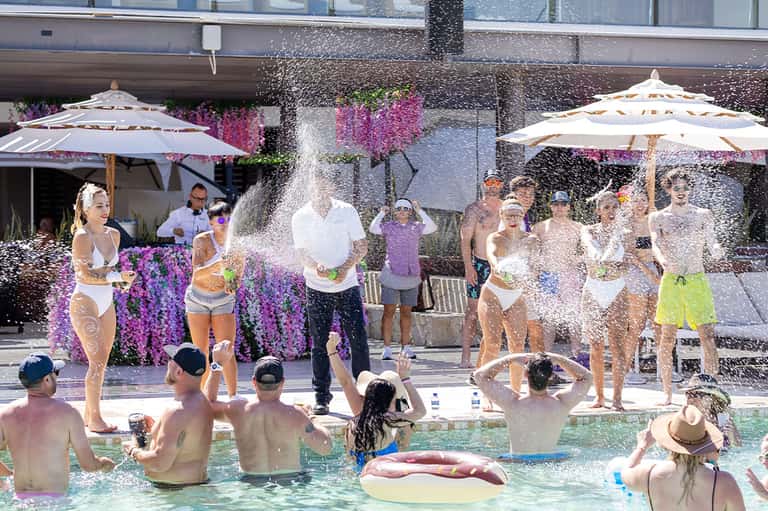 Pool party with guests in water, people standing under umbrellas, pink flowering plants visible