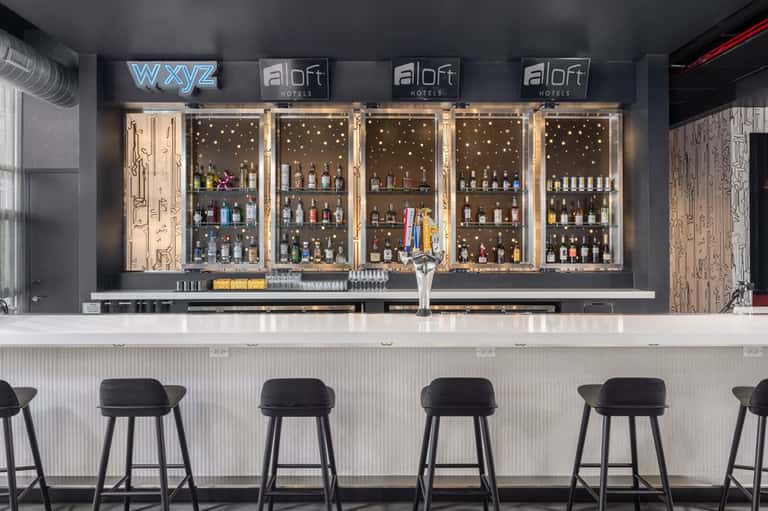 Modern hotel bar with illuminated shelving, black bar stools, and bottle display