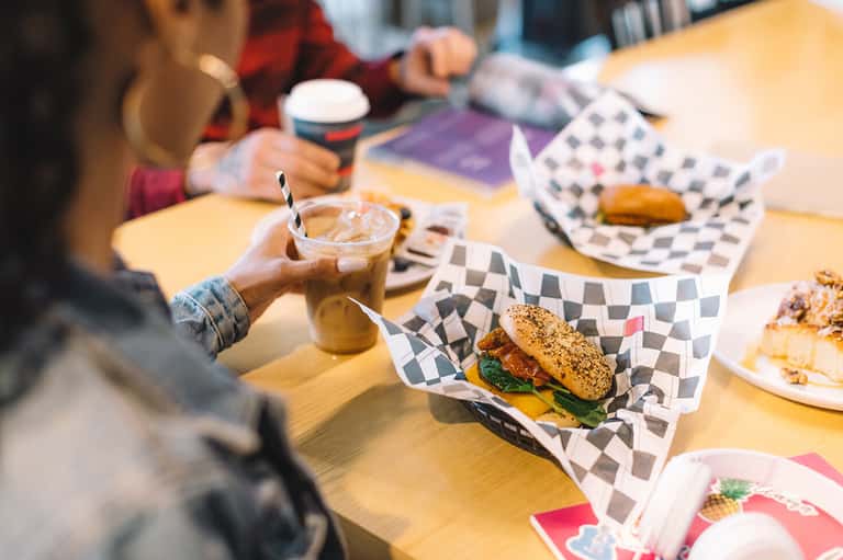Person enjoying iced coffee at casual indoor dining table with burgers served in checkered paper