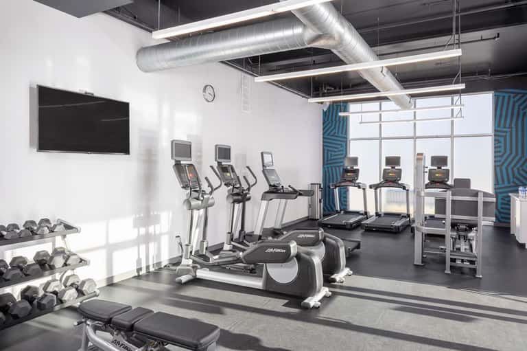 Modern fitness center with cardio equipment, weights, TV screen, and floor-to-ceiling windows