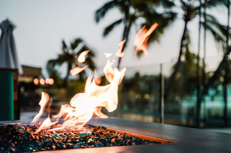 Outdoor fire pit with flames burning, palm trees visible in blurred background at dusk
